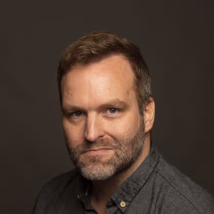 Colin Weldon, photographed in a dark grey shirt against a deep olive background